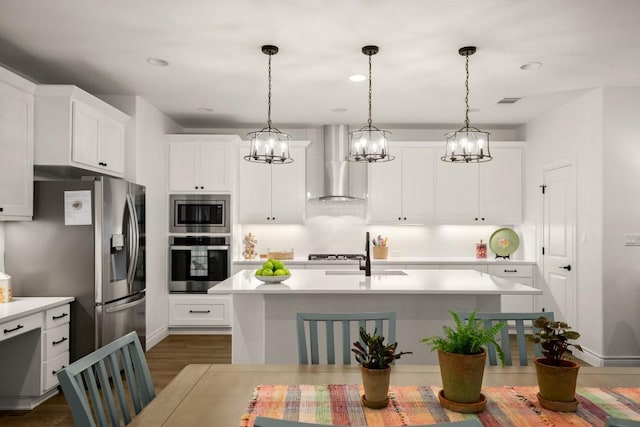 kitchen with appliances with stainless steel finishes, pendant lighting, white cabinets, a center island with sink, and wall chimney exhaust hood