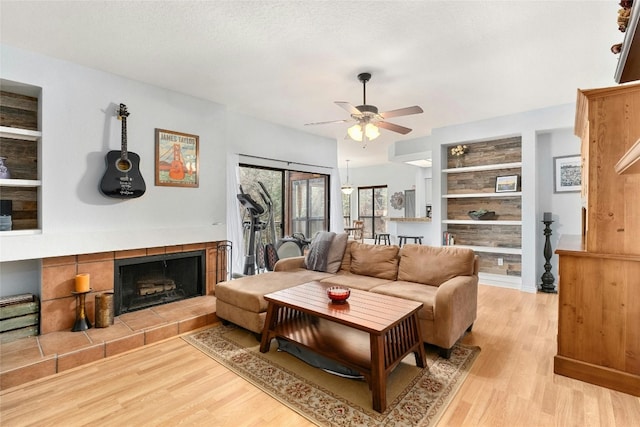 living room featuring built in features, a fireplace, ceiling fan, light hardwood / wood-style floors, and a textured ceiling