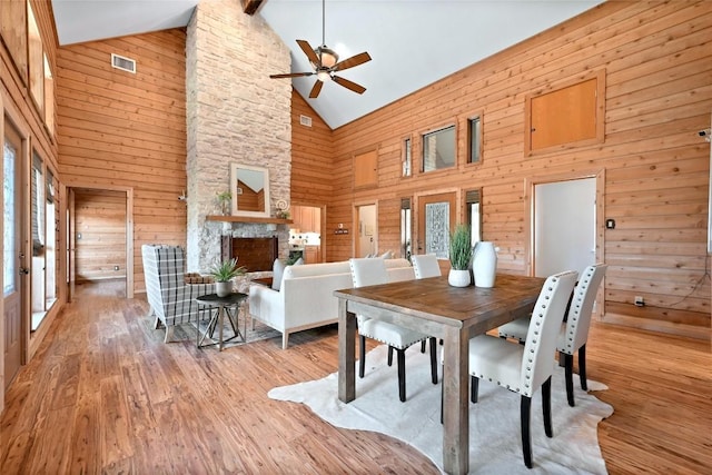 dining space featuring a fireplace, light wood-style floors, ceiling fan, wooden walls, and high vaulted ceiling