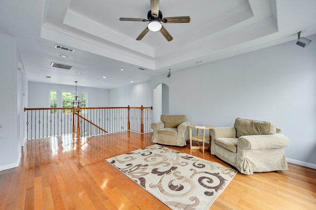 living area with a tray ceiling, wood-type flooring, ornamental molding, and ceiling fan with notable chandelier