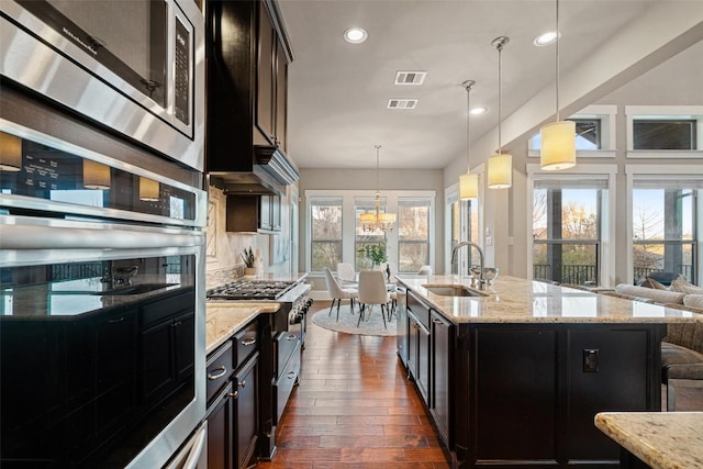 kitchen featuring sink, hanging light fixtures, stainless steel appliances, light stone counters, and a center island with sink