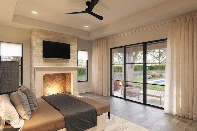 bedroom with access to outside, a tray ceiling, ceiling fan, a fireplace, and hardwood / wood-style floors