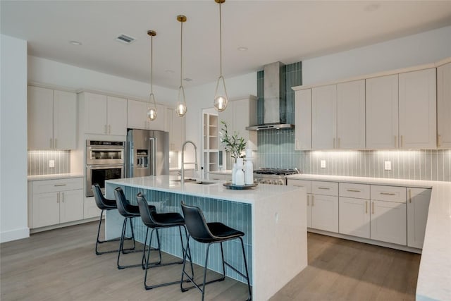 kitchen featuring sink, a center island with sink, appliances with stainless steel finishes, wall chimney range hood, and white cabinets