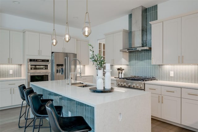 kitchen with pendant lighting, white cabinets, a kitchen island with sink, stainless steel appliances, and wall chimney range hood