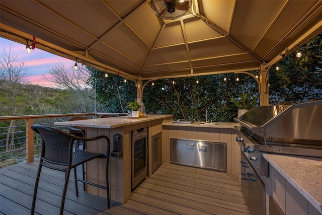 deck at dusk with wine cooler, exterior kitchen, an outdoor bar, grilling area, and a gazebo