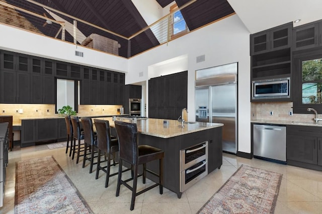 kitchen with sink, tasteful backsplash, built in appliances, an island with sink, and light stone countertops