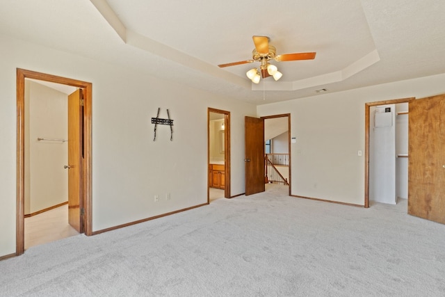 unfurnished bedroom with ensuite bathroom, a spacious closet, light colored carpet, a raised ceiling, and ceiling fan