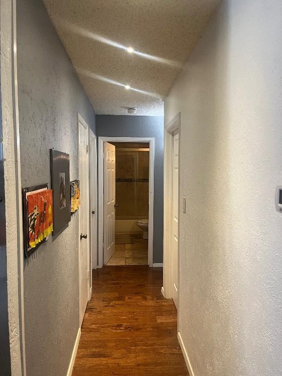 hall with a textured ceiling, a textured wall, wood finished floors, and baseboards