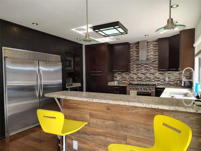 kitchen with built in appliances, a sink, wall chimney range hood, tasteful backsplash, and dark wood finished floors