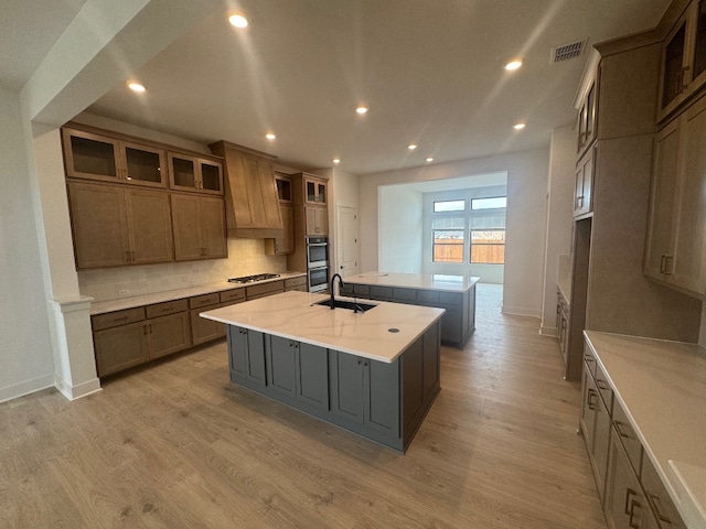 kitchen featuring sink, light hardwood / wood-style flooring, stainless steel appliances, light stone countertops, and a center island with sink