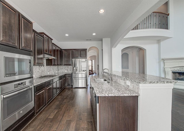 kitchen featuring sink, dark wood-type flooring, stainless steel appliances, dark brown cabinetry, and a center island with sink