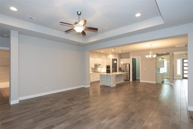 unfurnished living room featuring sink, dark wood-type flooring, ceiling fan with notable chandelier, and a tray ceiling