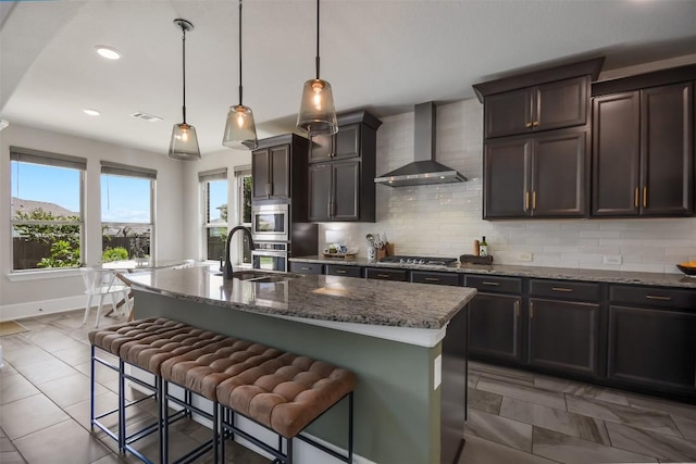 kitchen featuring wall chimney exhaust hood, a breakfast bar, decorative light fixtures, a center island with sink, and dark stone countertops