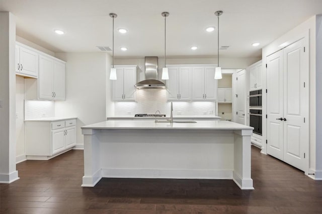 kitchen featuring white cabinetry, decorative light fixtures, a center island with sink, and wall chimney exhaust hood