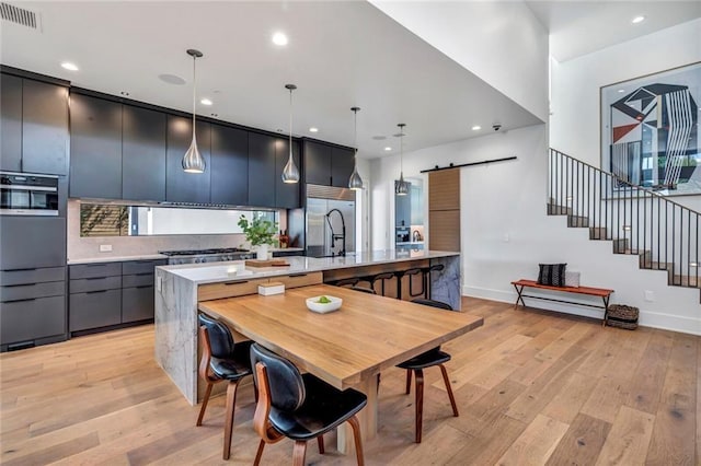 kitchen with decorative light fixtures, a large island with sink, stainless steel appliances, a barn door, and light hardwood / wood-style flooring