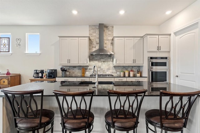 kitchen featuring appliances with stainless steel finishes, white cabinetry, an island with sink, a breakfast bar area, and wall chimney range hood