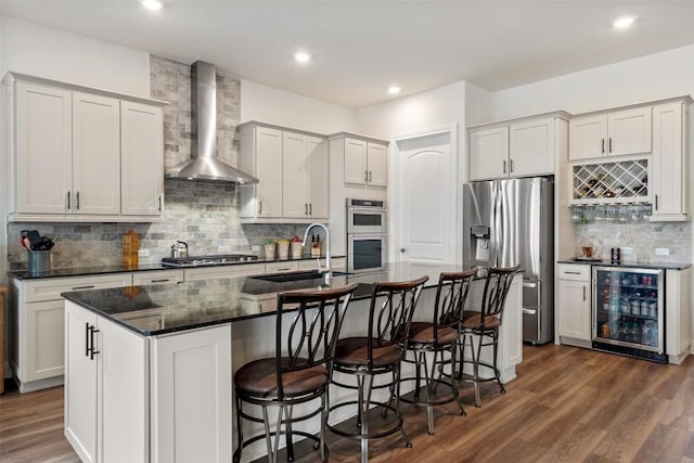 kitchen with sink, stainless steel appliances, beverage cooler, a kitchen island with sink, and wall chimney range hood