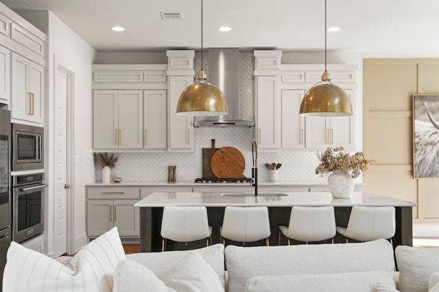 kitchen with a center island with sink, appliances with stainless steel finishes, wall chimney range hood, and decorative light fixtures