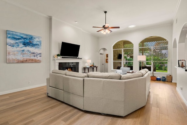 living room featuring ornamental molding, a fireplace, ceiling fan, arched walkways, and light wood finished floors