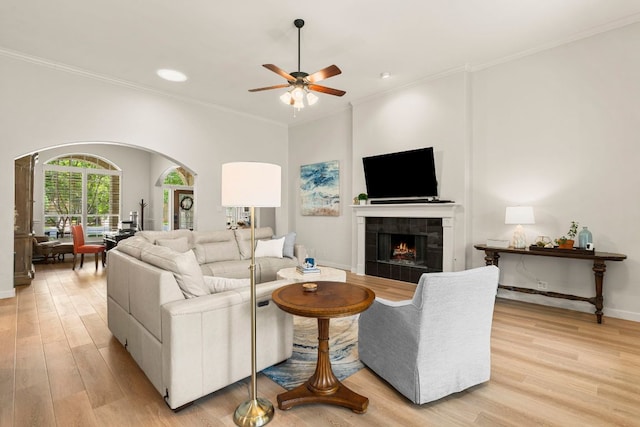 living area featuring light wood finished floors, arched walkways, a ceiling fan, a tile fireplace, and ornamental molding