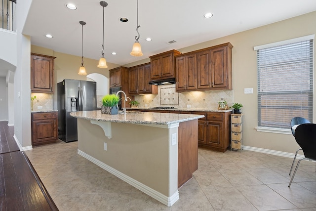 kitchen featuring stainless steel refrigerator with ice dispenser, gas stovetop, light stone counters, hanging light fixtures, and a center island with sink