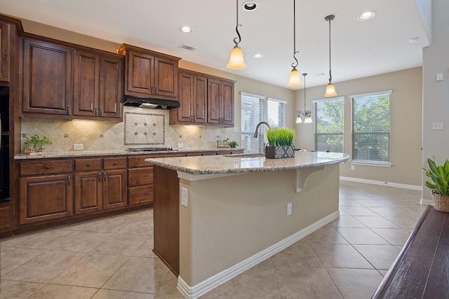 kitchen featuring light tile patterned floors, hanging light fixtures, light stone countertops, a center island with sink, and stainless steel gas stovetop