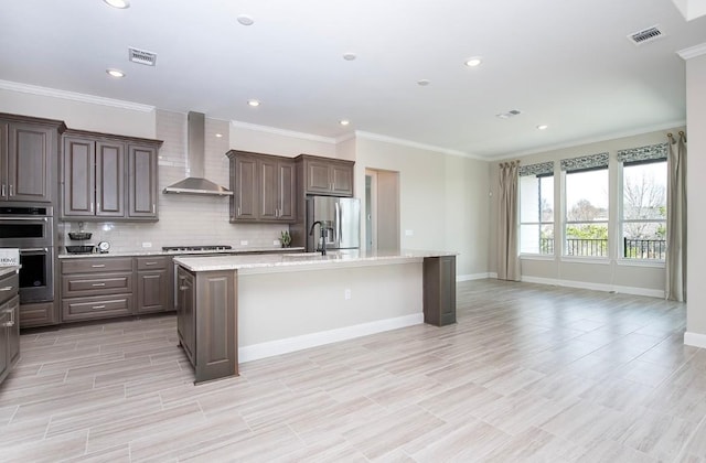 kitchen featuring dark brown cabinetry, a center island with sink, stainless steel appliances, decorative backsplash, and wall chimney range hood