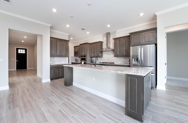 kitchen with dark brown cabinetry, appliances with stainless steel finishes, a center island with sink, and wall chimney range hood