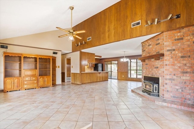 unfurnished living room featuring light tile patterned floors, wooden walls, high vaulted ceiling, ceiling fan with notable chandelier, and a wood stove