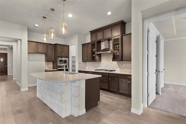 kitchen with pendant lighting, wall chimney range hood, appliances with stainless steel finishes, a kitchen island with sink, and tasteful backsplash