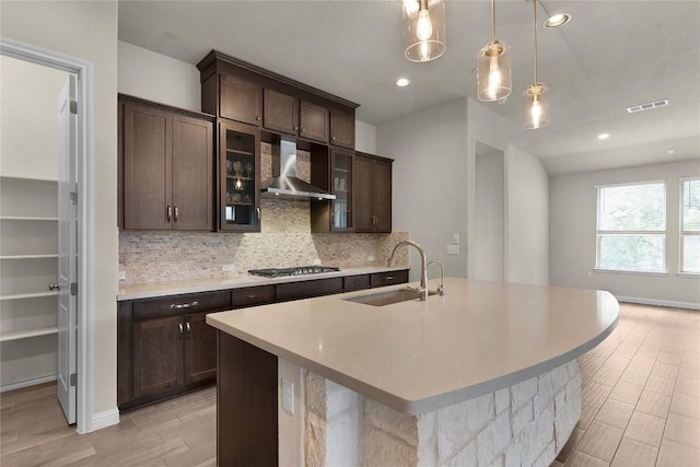 kitchen with decorative light fixtures, sink, decorative backsplash, a kitchen island with sink, and wall chimney range hood