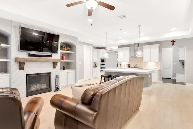 living room featuring sink, ceiling fan, a tray ceiling, a fireplace, and light wood-type flooring