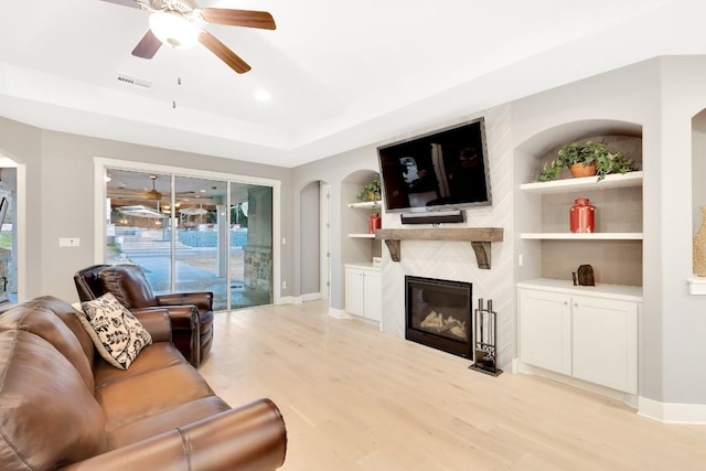 living room featuring built in shelves, light hardwood / wood-style flooring, a raised ceiling, ceiling fan, and a fireplace