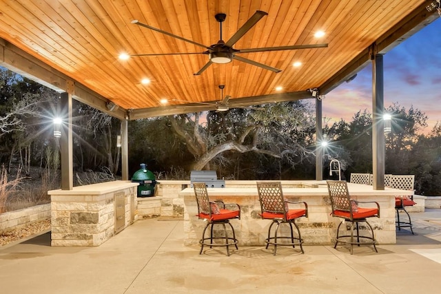 patio terrace at dusk featuring a bar, an outdoor kitchen, a grill, and ceiling fan