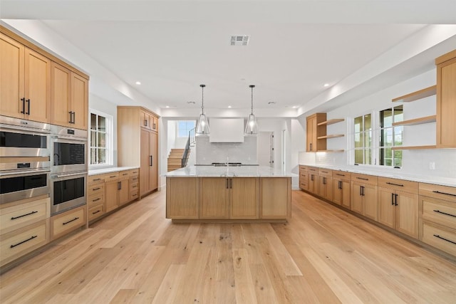 kitchen with hanging light fixtures, light hardwood / wood-style flooring, an island with sink, stainless steel double oven, and backsplash