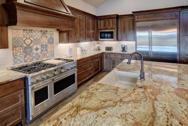 kitchen featuring sink, built in appliances, vaulted ceiling, custom range hood, and light stone countertops