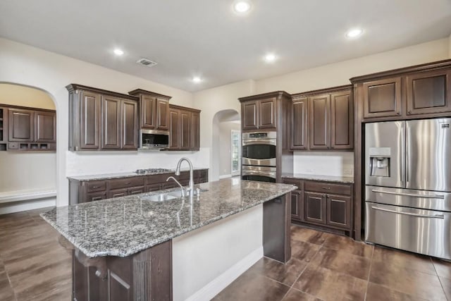 kitchen featuring dark brown cabinetry, sink, appliances with stainless steel finishes, dark stone counters, and a kitchen island with sink