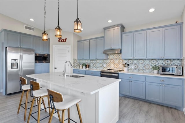 kitchen featuring sink, hanging light fixtures, appliances with stainless steel finishes, light stone countertops, and a kitchen island with sink