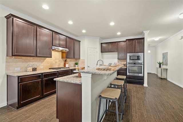 kitchen with light stone counters, tasteful backsplash, appliances with stainless steel finishes, dark hardwood / wood-style floors, and an island with sink