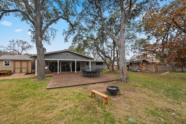 back of house featuring a storage shed, a lawn, a wooden deck, a playground, and a fire pit