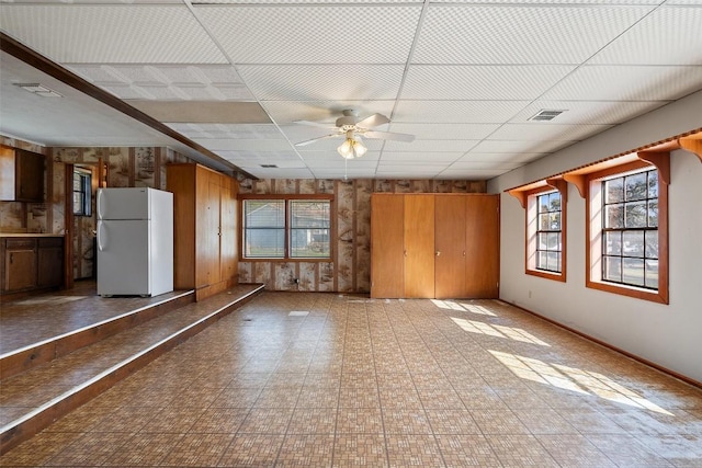 unfurnished living room featuring a drop ceiling, wooden walls, and ceiling fan