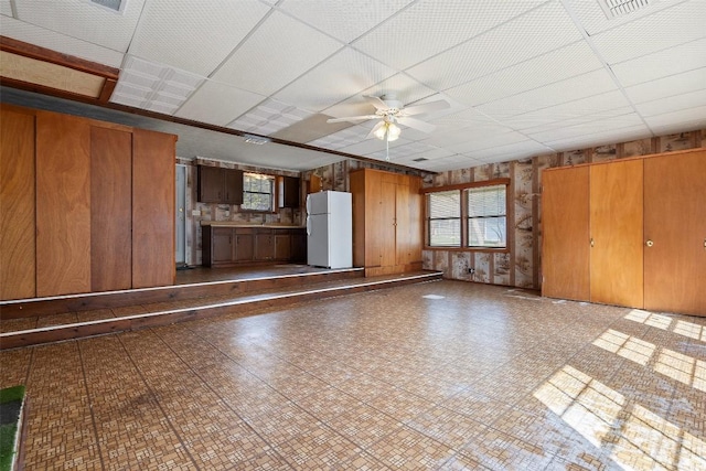 unfurnished living room featuring a drop ceiling, ceiling fan, and wood walls