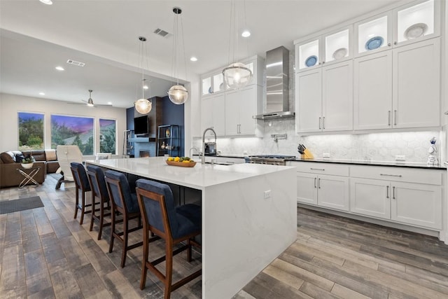 kitchen featuring dark wood-type flooring, white cabinetry, a center island with sink, pendant lighting, and wall chimney range hood