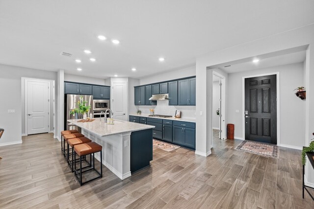 kitchen with appliances with stainless steel finishes, a kitchen bar, a kitchen island with sink, light stone counters, and light hardwood / wood-style floors