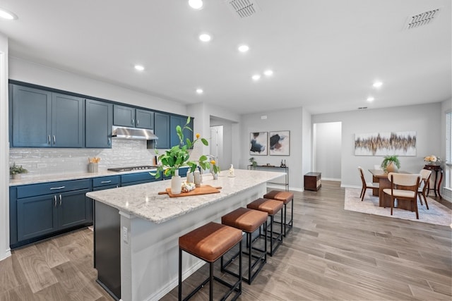 kitchen featuring a breakfast bar, decorative backsplash, stainless steel gas cooktop, a kitchen island with sink, and light wood-type flooring