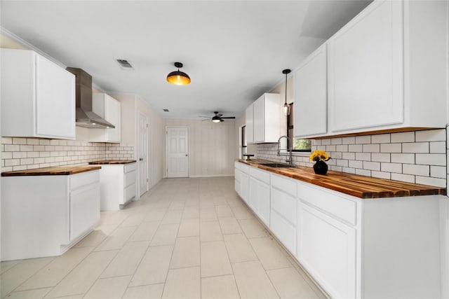 kitchen featuring wall chimney range hood, sink, ceiling fan, butcher block counters, and white cabinets