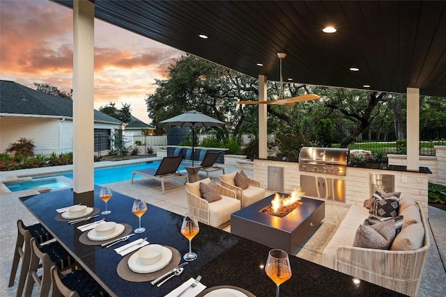 patio terrace at dusk featuring a grill, an outdoor kitchen, a fenced in pool, and an outdoor living space with a fire pit