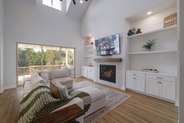 living room featuring baseboards, a glass covered fireplace, a towering ceiling, light wood-style flooring, and built in shelves