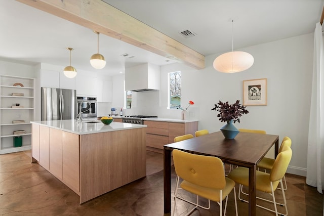 kitchen with wall chimney range hood, appliances with stainless steel finishes, white cabinets, a center island with sink, and beamed ceiling