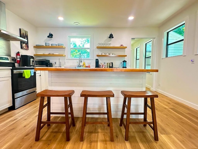 kitchen featuring wall chimney exhaust hood, stainless steel range with electric cooktop, light wood-style floors, white cabinetry, and open shelves